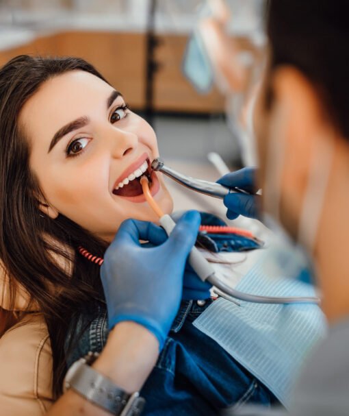 Young female patient visiting dentist office. Beautiful woman with healthy straight white teeth sitting at dental chair with open mouth during oral checkup while doctor working at teeth.